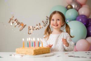 fofa pequeno menina golpes Fora velas em uma aniversário bolo às casa contra uma pano de fundo do balões. criança aniversário foto