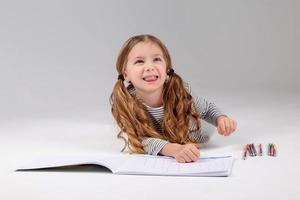 pequeno menina dentro uma listrado vestir desenha dentro a álbum deitado em a chão. criança desenvolvimento. estilo de vida. pré escola e escola Educação. espaço para texto. Alto qualidade foto