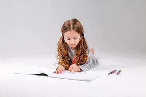 pequeno menina dentro uma listrado vestir desenha dentro a álbum deitado em a chão. criança desenvolvimento. estilo de vida. pré escola e escola Educação. espaço para texto. Alto qualidade foto