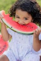 uma pequeno moreno menina com encaracolado cabelo dentro uma pálido Rosa vestir come uma Melancia em a grama. piquenique dentro a parque. feliz infância. espaço para texto. Alto qualidade foto