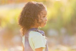 retrato pequeno feliz brincalhão criança pequena moreno menina dentro uma jeans vestido de verão em pé dentro a jardim em uma ensolarado dia. caminhando dentro a fresco ar. conceito do uma feliz infância. espaço para texto. Alto qualidade foto