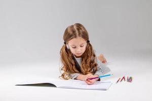 pequeno menina dentro uma listrado vestir desenha dentro a álbum deitado em a chão. criança desenvolvimento. estilo de vida. pré escola e escola Educação. espaço para texto. Alto qualidade foto