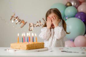 fofa pequeno menina golpes Fora velas em uma aniversário bolo às casa contra uma pano de fundo do balões. criança aniversário foto