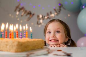 fofa pequeno menina golpes Fora velas em uma aniversário bolo às casa contra uma pano de fundo do balões. criança aniversário foto