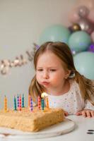 fofa pequeno menina golpes Fora velas em uma aniversário bolo às casa contra uma pano de fundo do balões. criança aniversário foto