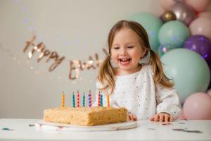 fofa pequeno menina golpes Fora velas em uma aniversário bolo às casa contra uma pano de fundo do balões. criança aniversário foto