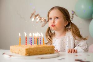 fofa pequeno menina golpes Fora velas em uma aniversário bolo às casa contra uma pano de fundo do balões. criança aniversário foto