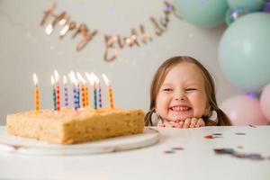 fofa pequeno menina golpes Fora velas em uma aniversário bolo às casa contra uma pano de fundo do balões. criança aniversário foto