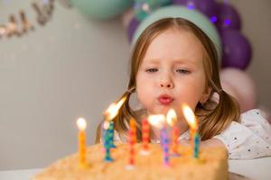 fofa pequeno menina golpes Fora velas em uma aniversário bolo às casa contra uma pano de fundo do balões. criança aniversário foto