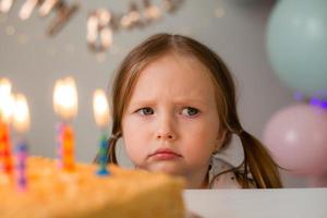 fofa pequeno menina golpes Fora velas em uma aniversário bolo às casa contra uma pano de fundo do balões. criança aniversário foto