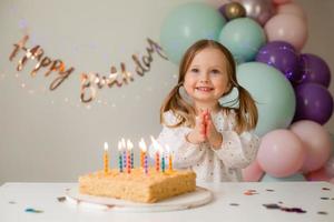 fofa pequeno menina golpes Fora velas em uma aniversário bolo às casa contra uma pano de fundo do balões. criança aniversário foto