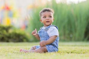 uma fofa pele escura bebê dentro uma jeans macacão é sentado em a grama. feliz infância. espaço para texto. Alto qualidade foto
