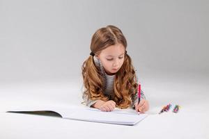 pequeno menina dentro uma listrado vestir desenha dentro a álbum deitado em a chão. criança desenvolvimento. estilo de vida. pré escola e escola Educação. espaço para texto. Alto qualidade foto