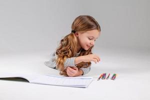 pequeno menina dentro uma listrado vestir desenha dentro a álbum deitado em a chão. criança desenvolvimento. estilo de vida. pré escola e escola Educação. espaço para texto. Alto qualidade foto