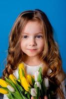retrato do uma encantador sorridente pequeno menina com uma ramalhete do tulipas dentro dela mãos. estilo de vida. fresco flores internacional mulheres dia. espaço para texto. Alto qualidade foto
