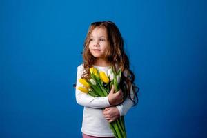 retrato do uma encantador sorridente pequeno menina com uma ramalhete do tulipas dentro dela mãos. estilo de vida. fresco flores internacional mulheres dia. espaço para texto. Alto qualidade foto