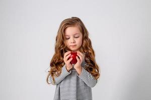 fofa pequeno menina com cachos dentro uma listrado vestir é segurando uma grande vermelho maçã dentro dela mãos. azul fundo. útil produtos para crianças. saudável lanche. espaço para texto. Alto qualidade foto