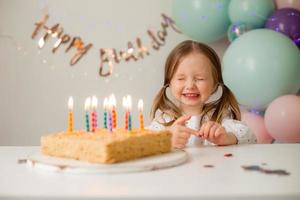 fofa pequeno menina golpes Fora velas em uma aniversário bolo às casa contra uma pano de fundo do balões. criança aniversário foto