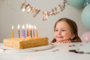 fofa pequeno menina golpes Fora velas em uma aniversário bolo às casa contra uma pano de fundo do balões. criança aniversário foto