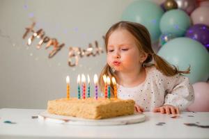 fofa pequeno menina golpes Fora velas em uma aniversário bolo às casa contra uma pano de fundo do balões. criança aniversário foto