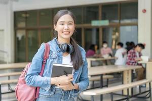 bela estudante mulher asiática com mochila e livros ao ar livre. menina sorriso feliz carregando um monte de livro no campus da faculdade. retrato feminino na universidade internacional da ásia. educação, estudo, escola foto