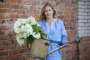 uma lindo idosos mulher dentro a bordado camisa detém uma retro rega pode com flores a conceito do floricultura e jardinagem Como uma passatempo. foto