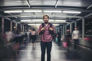 bonito homem barbudo com mochila em pé na rua enquanto viaja à noite foto