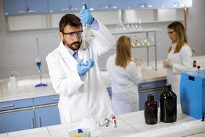 jovem pesquisador em roupas de trabalho de proteção em pé no laboratório e analisando frasco com líquido