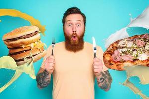 homem com tatuagens é pronto para comer sanduíche e pizza com talheres dentro mão. ciano fundo foto