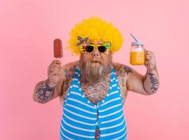 gordo surpreso homem com barba e peruca come uma picolé e bebidas uma suco fruta foto