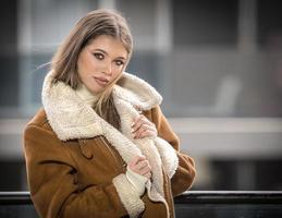 linda sorridente menina dentro à moda roupas posando em moderno sacada com surpreendente Visão em cidade .retrato do uma à moda elegante Loiras Adolescência com pele casaco com ampla coleiras vestindo dentro sacada foto