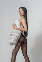 mulher sensual em meia-calça preta, pelo curto branco e pernas longas posando desafiadora na frente do fundo. cena clássica de boudoir. foto erótica de estúdio. garota atraente com salto alto na cadeira alta