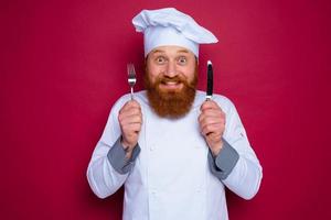 feliz chefe de cozinha com barba e vermelho avental detém talheres dentro mão foto