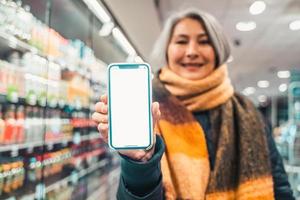 Senior mulher às a supermercado mostra a tela do dela Smartphone foto