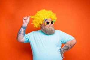 gordo feliz homem com barba, tatuagens e oculos de sol tem Diversão com a amarelo peruca foto