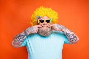 gordo feliz homem com barba, tatuagens e oculos de sol tem Diversão com a amarelo peruca foto