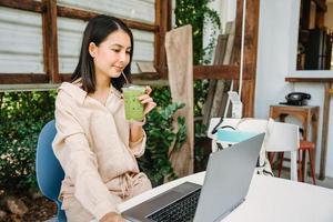 jovem ásia mulher bebendo gelado verde chá e trabalhando com computador portátil às escrivaninha dentro quintal às café fazer compras foto