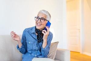 linda mulher idosa caucasiana conversando por telefone com seu velho amigo usando dispositivo eletrônico, discutindo as últimas notícias com um sorriso fofo no rosto, sentado em casa foto