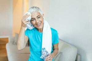 Senior mulher segurando plástico garrafa do água, limpando suor com uma toalha, Exausta depois de a diariamente treinamento. idosos mulher levando uma pausa enquanto exercício às lar. sentado em a sofá foto