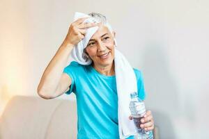 Senior mulher segurando plástico garrafa do água, limpando suor com uma toalha, Exausta depois de a diariamente treinamento. idosos mulher levando uma pausa enquanto exercício às lar. foto