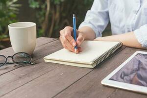 mão mulher escrevendo caderno em madeira mesa com copo café e tábua. foto