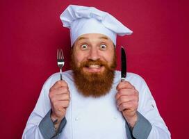 feliz chefe de cozinha com barba e vermelho avental detém talheres dentro mão foto