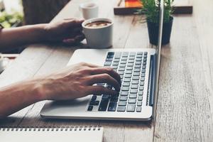 o negócio homem digitando computador portátil e segurando café copo em madeira mesa. foto