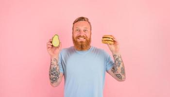 feliz homem com barba e tatuagens é indeciso E se para comer a abacate ou a Hamburger foto