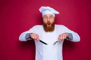 espantado chefe de cozinha com barba e vermelho avental detém talheres dentro mão foto