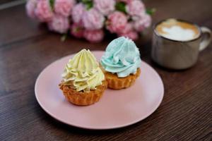 uma Rosa prato com aniversário bolinhos senta em uma de madeira mesa. uma bege copo do cappuccino senta em a mesa. perto uma prato do bolos mentira lindo Rosa e branco flores foto