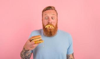com fome homem com barba e tatuagens come uma sandwitch com Hamburger e batatas foto