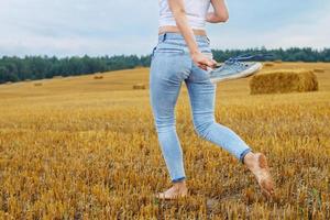 descalço menina com tênis dentro mão caminhando dentro a agrícola campo com palheiro e fardos foto