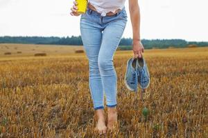 descalço menina com tênis e cartão copo com café dentro mão ficar de pé dentro a agrícola campo foto