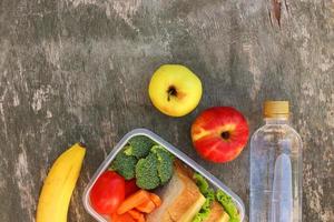 sanduíches, frutas e legumes na caixa de comida, água em fundo de madeira velho. vista do topo. postura plana. foto